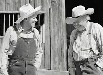 black and white photo of two old farmers standing in front of a barn and laughing