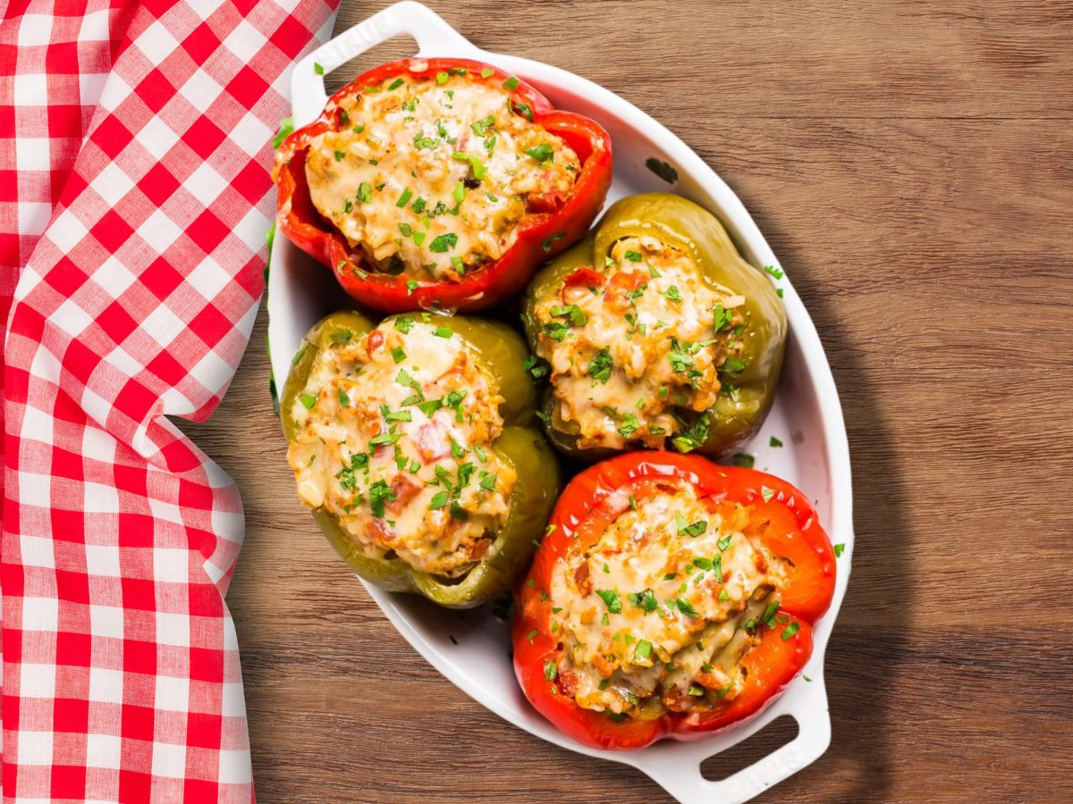 two red and two green stuffed bell peppers in a white serving dish on a brown cutting board with a red gingham table cloth beside it